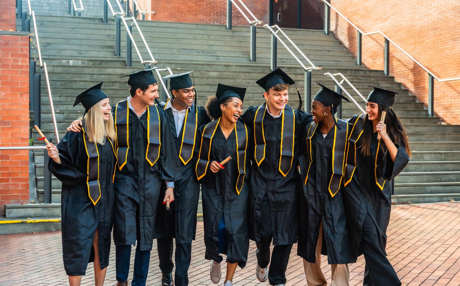 Group of smiling graduates in caps and gowns walking together and holding diplomas outdoors.